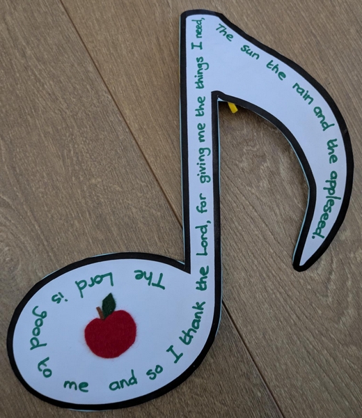 Cutout musical note with a felt 2-D apple stuck on and written text: The Lord is good to me and so I thank the Lord for giving me the things I need: the sun, the rain and the appleseed.