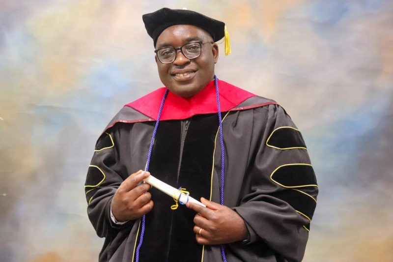 Dr Gilbert Esambe in his graduation cap and gown, holding his degree certificate.