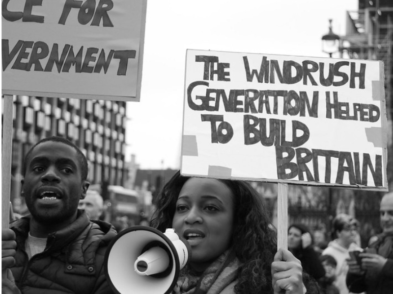 A crowd protesting against the treatment of the Windrush generation by the government. Centre, a woman holds a placard saying "The Windrush Generation helped to build Britain".